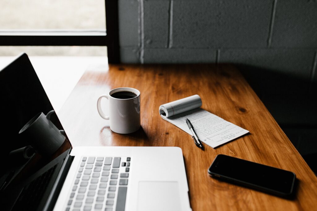 laptop, coffee mug, phone, and pen and paper sitting on wood table